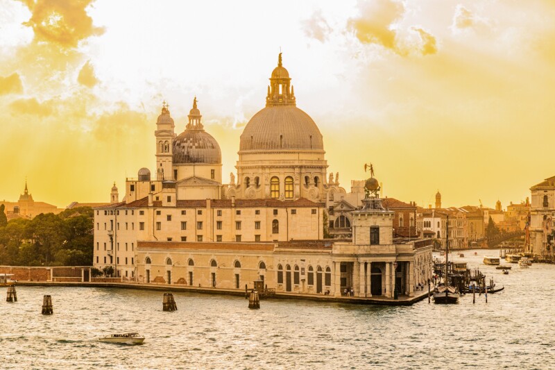 A domed building jutting out onto the water backed by a yellow cloudy sky in Venice.