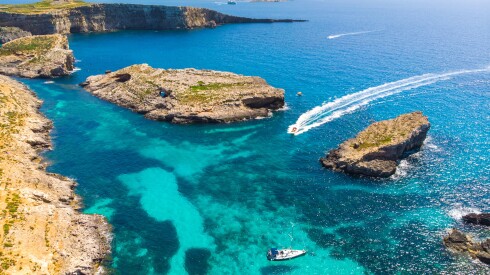 A high  view of a few speed boats on the bright blue ocean near the rocky coast line of Malta's Comino Island.