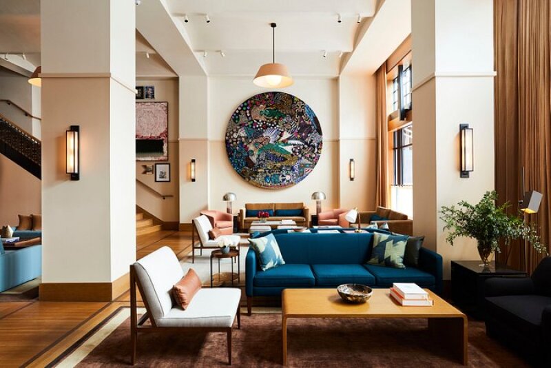 The lobby consists of many coffee tables surrounded by royal blue and cream white industrial furniture. There is an artistic centerpiece in the lobby, flashing peacock-like colors. Stairs on the left.