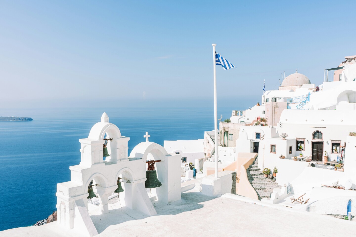 White buildings with bells and domes and a Greek flag on a flagpole overlooking the sea 