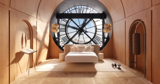 A bedroom inside Paris' Musée d'Orsay with a modern white and beige bed, a book stand and a huge clock in a window in the background