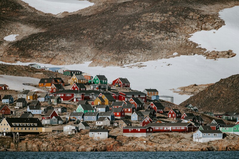 Small red, blue, green, and yellow houses line a rocky Greenland coastline, with sheet of snow on hill in background