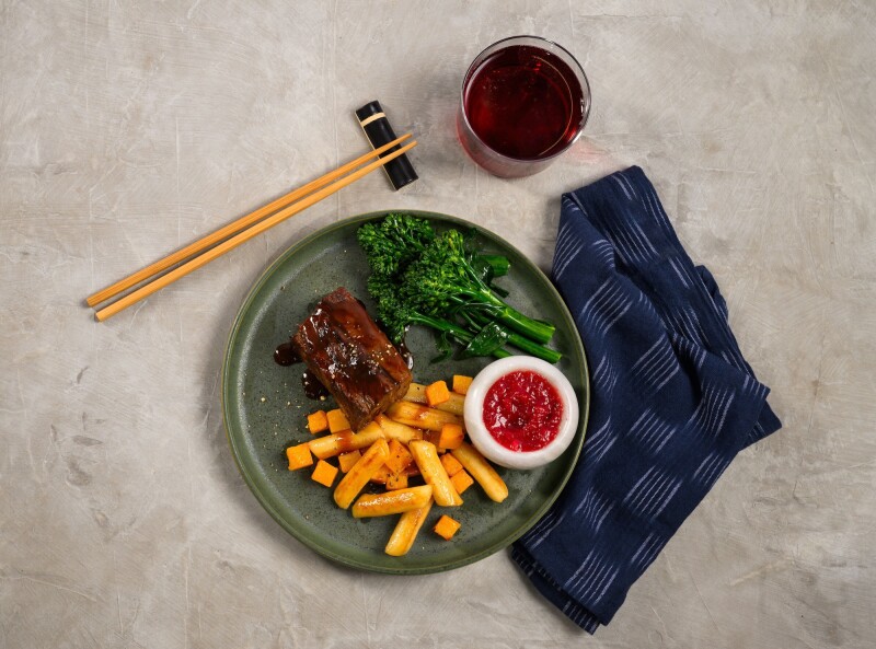 Alaska Airlines inflight meal is a piece of braised meat, orange vegetables, and green vegetables on a dark green dish with a white cup of red sauce. A blue napkin is under the plate.