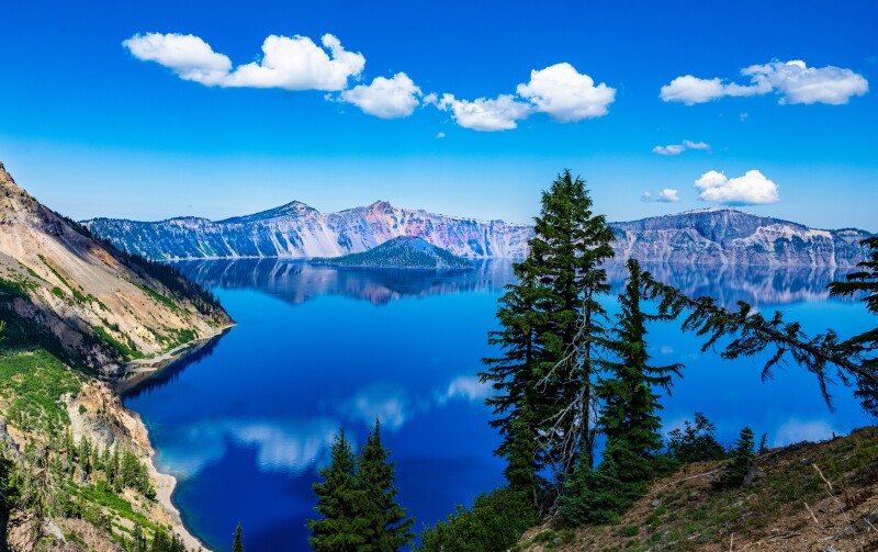 A blue lake surrouded by mountains, beneath a blue sky with a few white clouds, with a couple of evergreen trees in the foreground