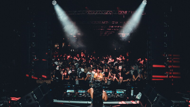 View from behind a DJ with long dark hair playing for a crowded dark room with two white spotlights and red accent lights at one of the Movement Music Festival’s after parties