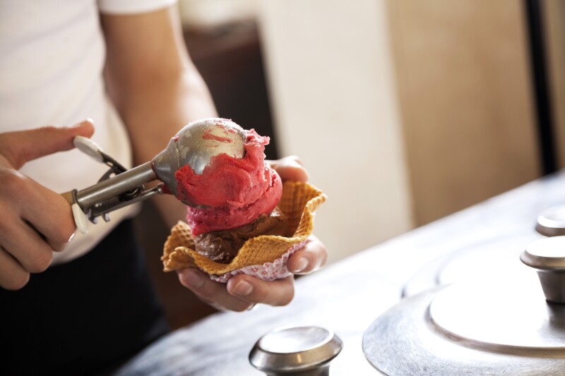 Hands of server with silver scoop of red gelato atop scoop of chocolate in wavy wafer