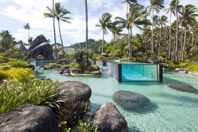 Laucala Island features both a lagoon pool and a raised pool with glass walls; palm trees surround both.