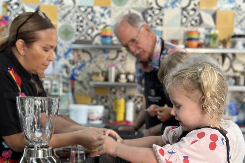 A young girl in the foreground using her hands to make something during a cooking lesson, with another child next to her and an elderly man in the background, while a woman stands opposite them in a kitchen
