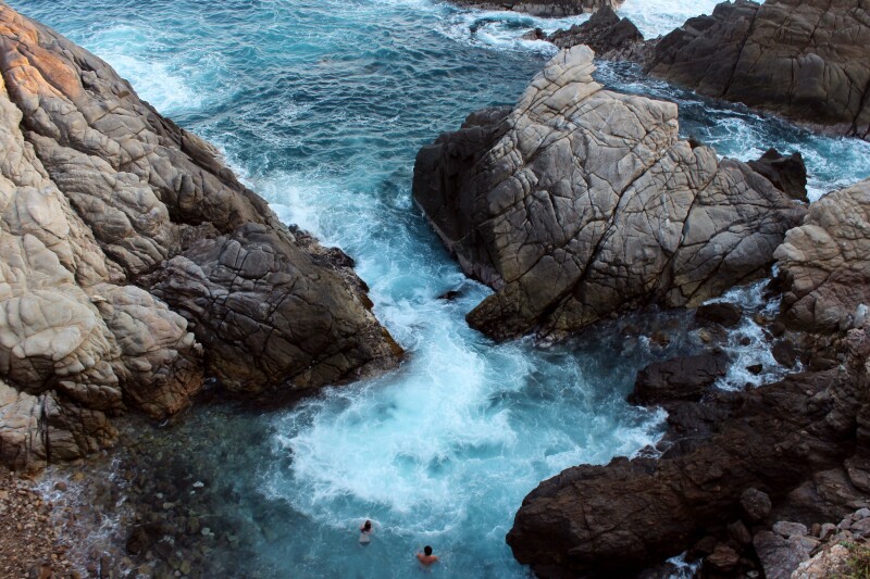 Overhead view of waves crashing between large boulders with two people in the surf