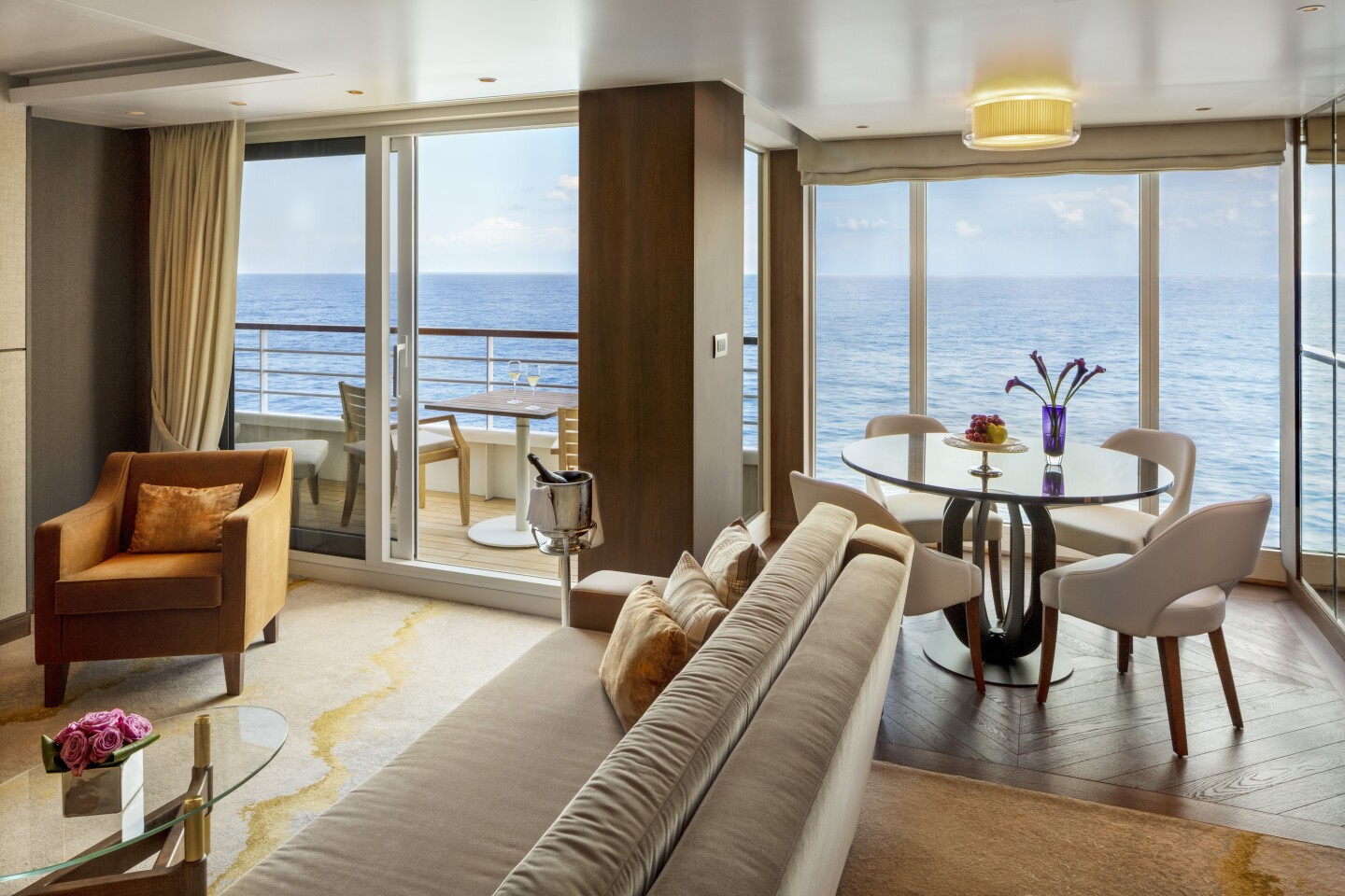View of the living area and a dining table in the Crystal Symphony penthouse suite with large windows and a balcony looking out at the sea
