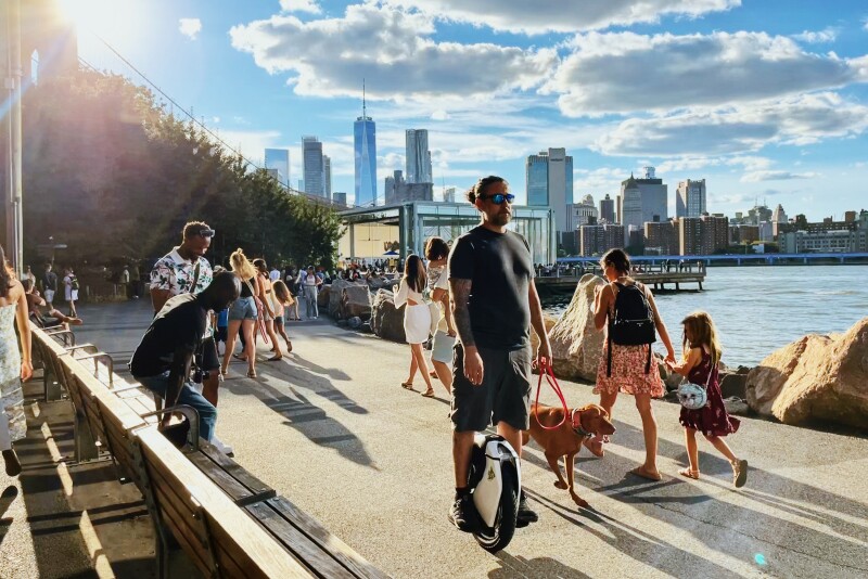 People on paved path in Brooklyn Bridge Park, with person on unicycle in front and Manhattan skyline in background