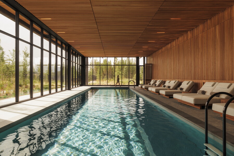A person walking along One&Only Moonlight Basin's indoor pool with wood paneling and modern loungers lining one side of the pool and floor-to-ceiling windows showcasing the surrounding trees along the other two sides
