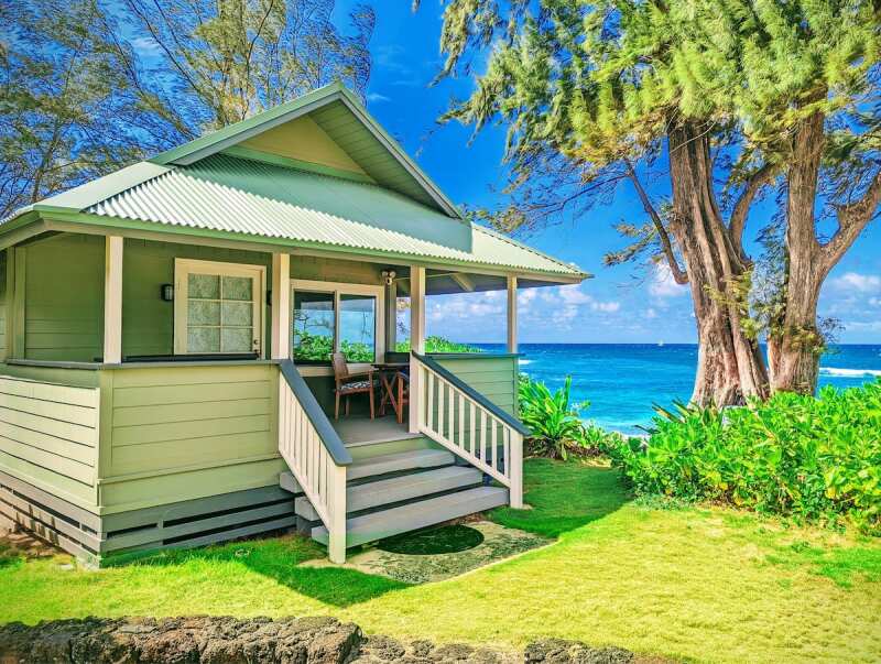 A small wooden cottage with a porch and short staircase next to a tree, with ocean in background
