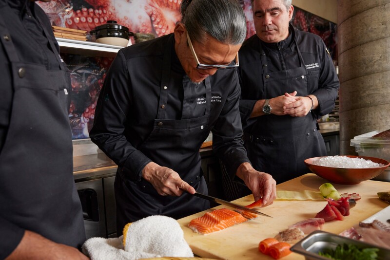 Chef Masaharu Morimoto in a black shirt and apron slices fresh salmon while other cooks watch