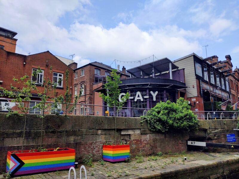 Manchester's Gay Village neighborhood, with restaurants along a street behind a canal. Trees are planted in pride-striped planter boxes, and a purple restaurant has "GAY" spelled out in big letters.