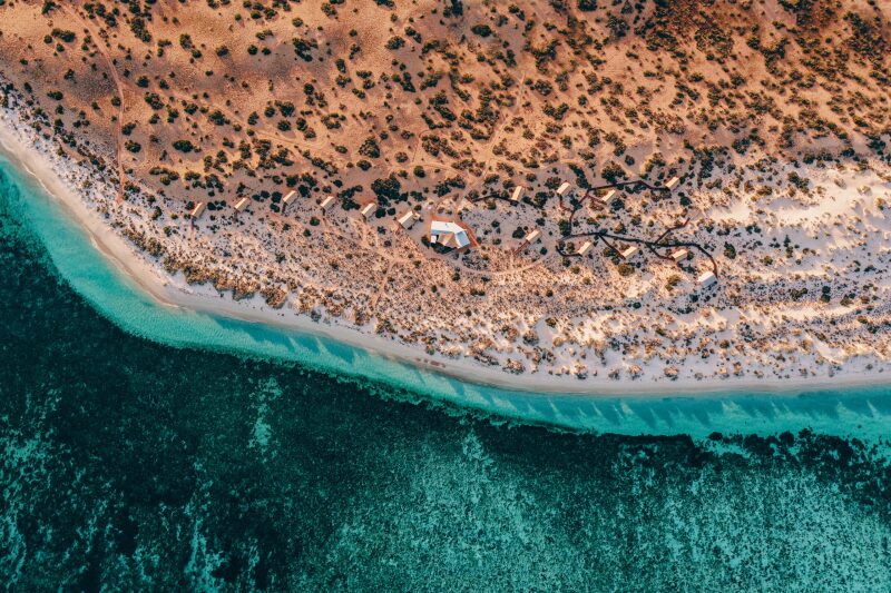 Aerial view of a beach with about 15 beige tents scattered around a bigger tent, the blue water filled with coral and vegetation