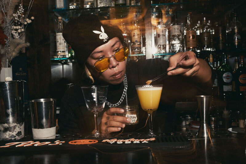 Bartender Katrina Sobredilla carefully garnishes frothy yellow cocktail at dimly lit bar, shelves of spirits visible behind her.