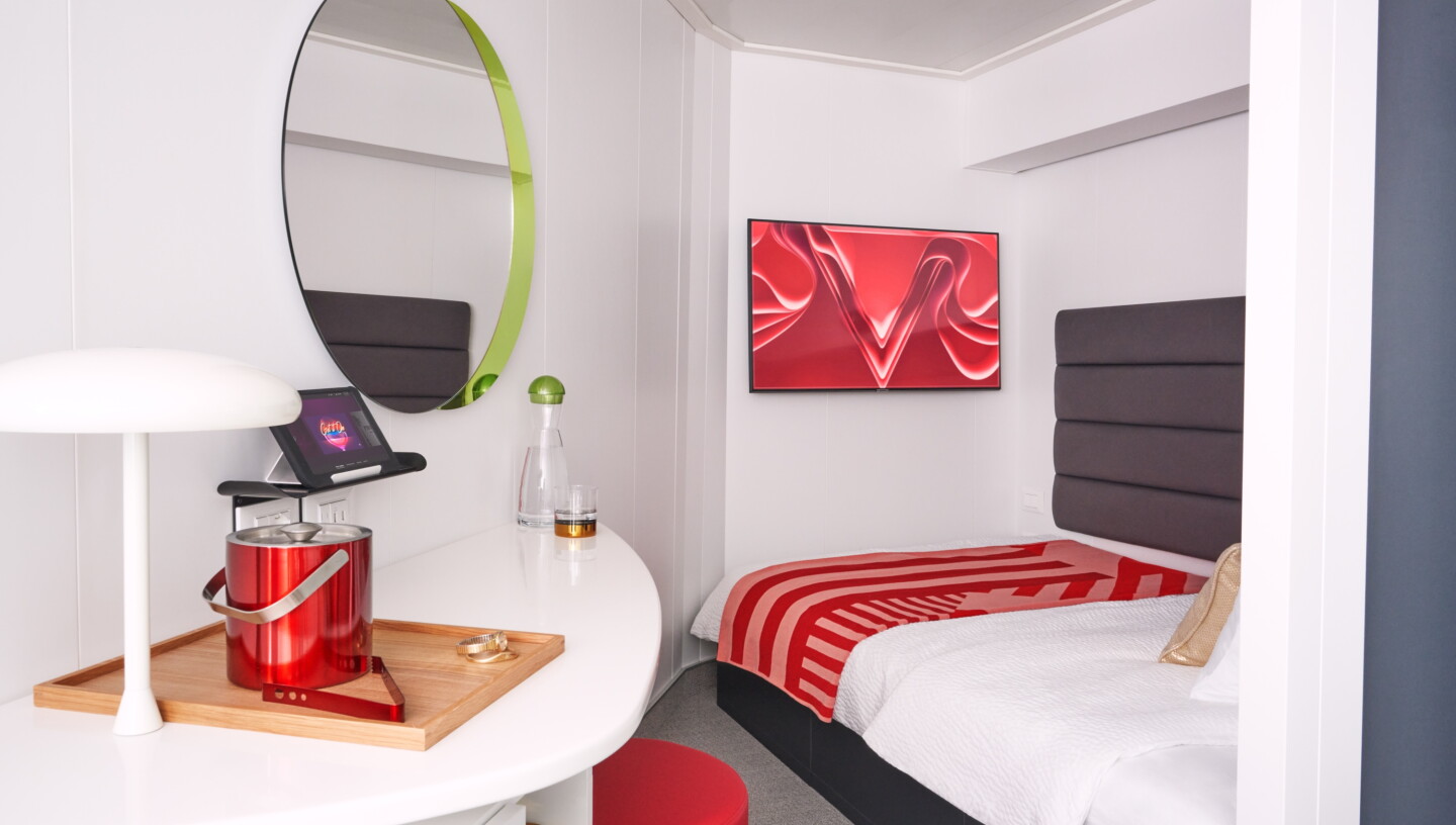 A solo cabin on a Virgin Voyages cruise ship, with single bed and desk, with red decor accents