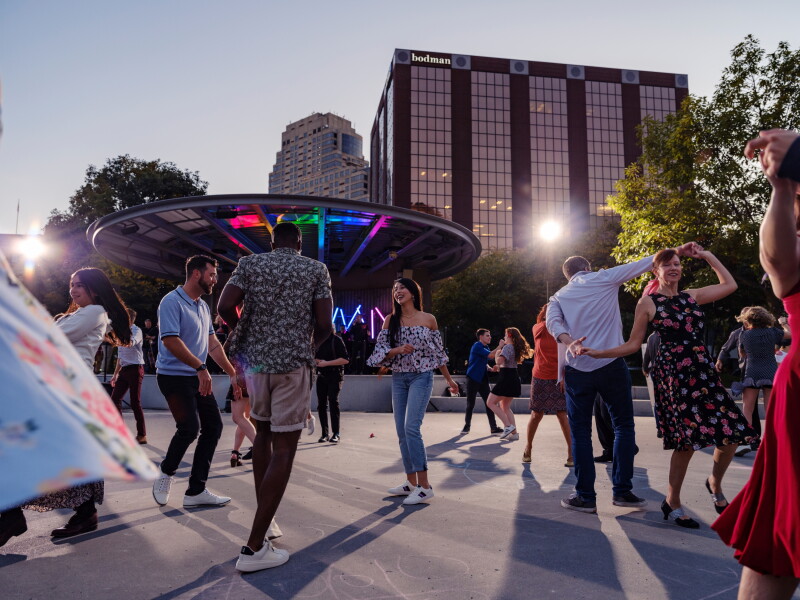 A dozen people swing dancing outside in Rosa Parks Circle, with large buildings and a disc-like installation with rainbow lights behind them