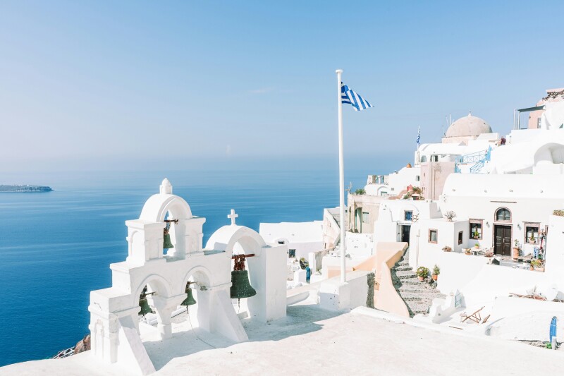 White buildings with bells and domes and a Greek flag on a flagpole overlooking the sea