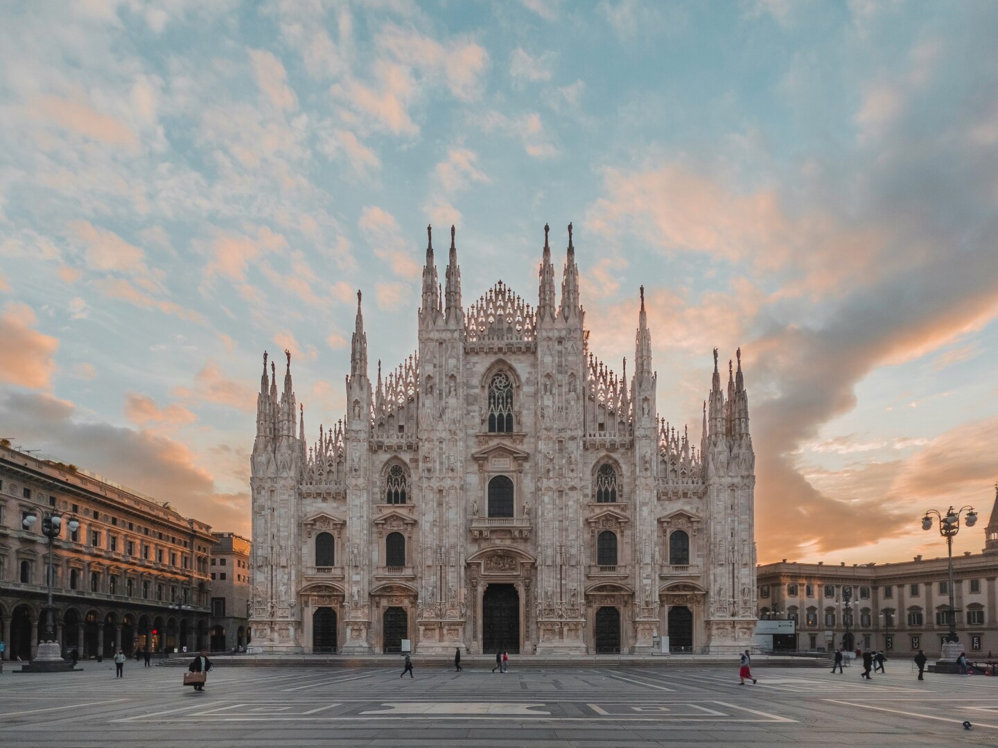 A view of the facade of the iconic Duomo di Milano cathedral in Milan viewed at sunrise with a small group of people walking around the square