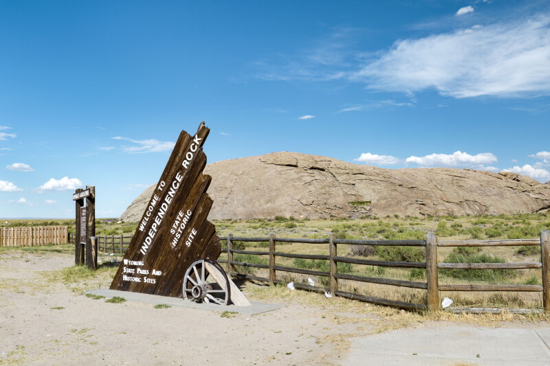 The wooden sign of Independence Rock State Historic Site in Wyoming, shaped like a wagon sticking out of the ground, with dry prairie and mountains beyond it
