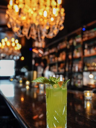 A green drink in a highball glass garnished with basil sits on the end of a dark wood bar with large chandelier overhead at Death and Taxes in Reno