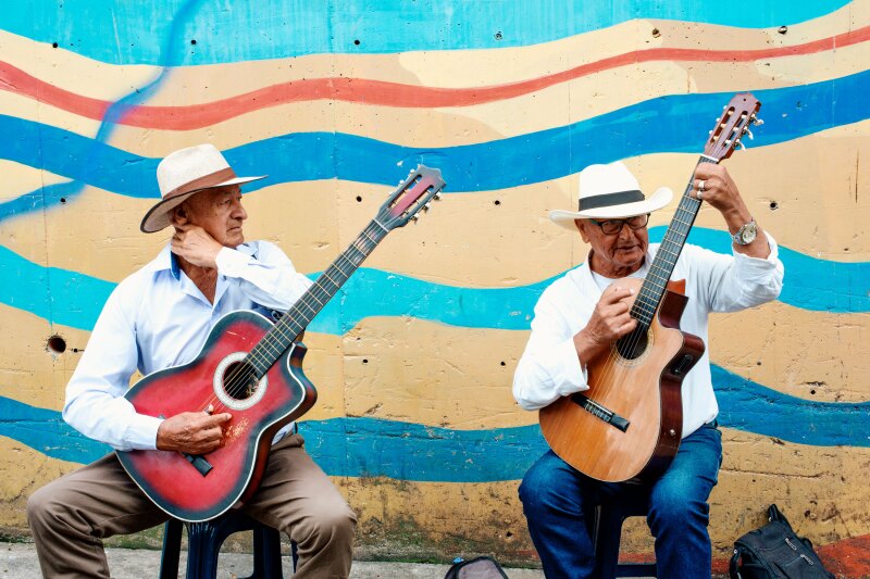 Two men wearing brimmed hats and white shirts sit on stools in front of a blue, yellow, and orange street mural and play their guitars, "Tour Comuna 13", Comuna 13, Medellín