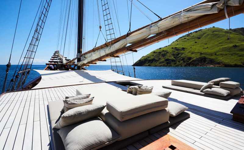 The yacht style deck on the Amandira sailing ship with padded loungers facing the front of the ship, and a large mast with a rolled up sail, with a lush green island in the background in Indonesia