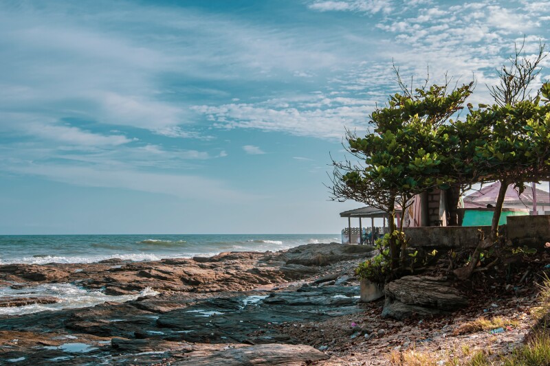 A rocky coastline with small waves breaking in Accra, Ghana, with some trees and structures along the coast