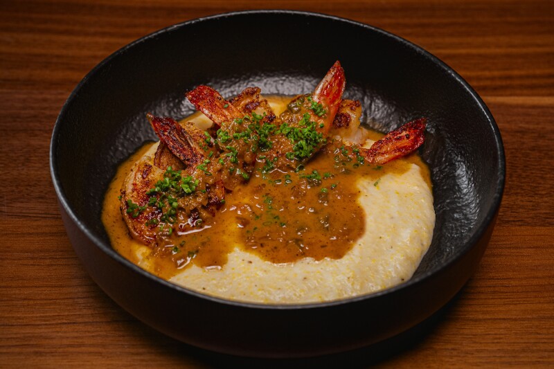 Overhead view of shrimp and grits from chef Kwame Onwuachi, with gravy and chives and served in shallow black bowl