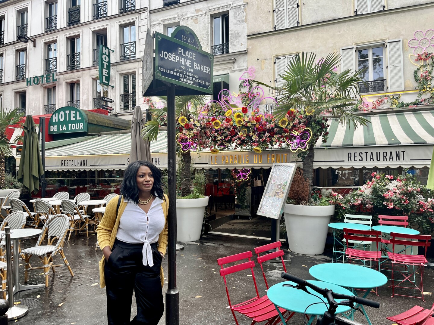 The writer leans against a Place Josephine Baker sign in Montaparnasse, Paris with cafe awnings and colorful cafe tables and chairs on the sidewalk around her