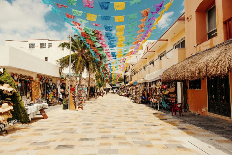 A view down the middle of a stone-paved market street lined with shops displaying wares in front and colorful flags covering the street overhead