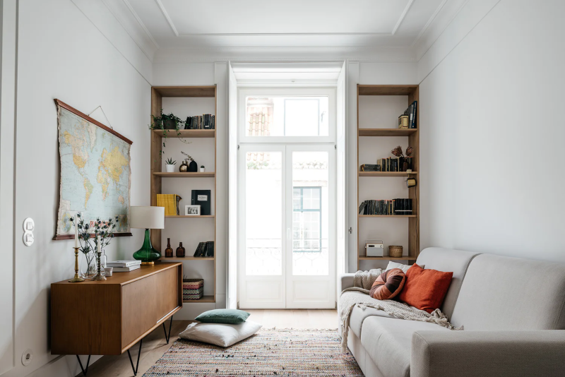 Interior of a Lisboans serviced apartment, with a pale gray sofa, built-in residential-style bookshelves surrounding tall window, and large map of world on wall