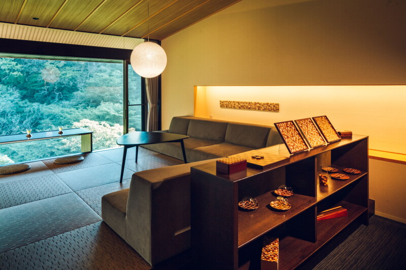 A guest room with an L-shaped sofa, a credenza, tatami mats, and a floor-to-ceiling window overlooking the forest