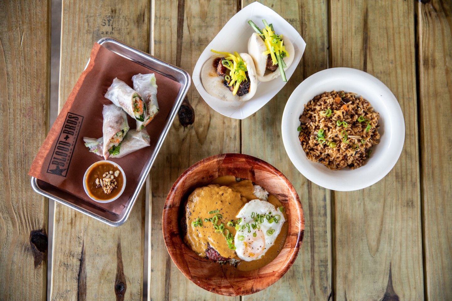Overhead view of spring rolls, brisket fried rice, loco moco, and brisket stuffed bao at Blood Bros. BBQ, on wooden table top