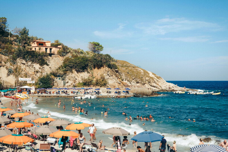 Busy Mediterranean beach, with swimmers in water, rows of orange umbrellas on sand, and rocky hillside with a terra-cotta villa and pine trees in left background