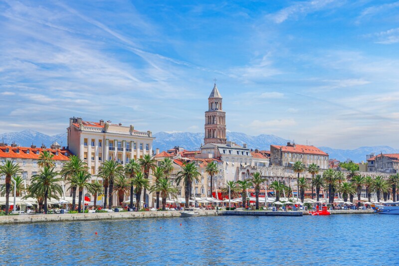 A palm-lined waterfront in Split, Croatia, with historic architecture, orange tile roofs, and snowcapped mountains in the background