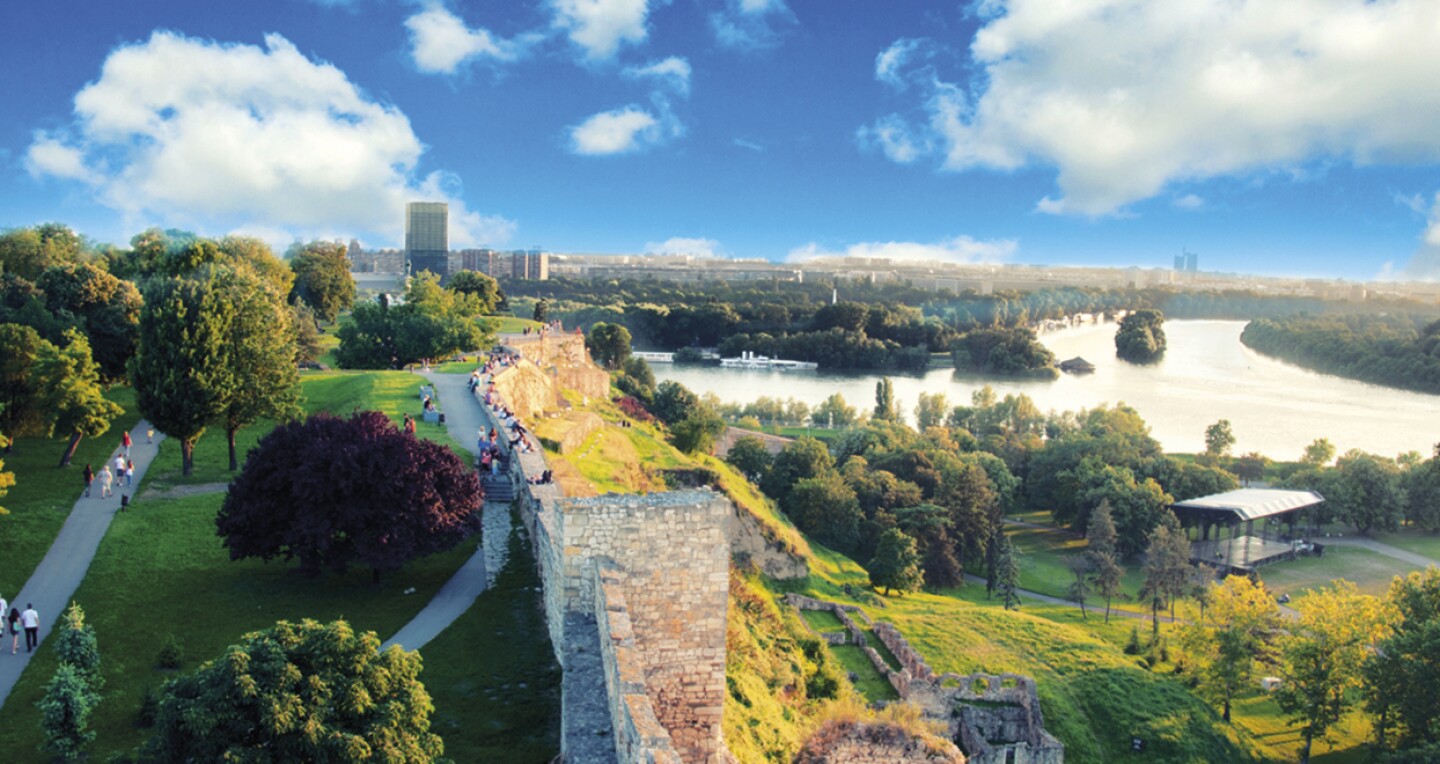 People sit along a wall overlooking a large river. A city is seen in the distance.