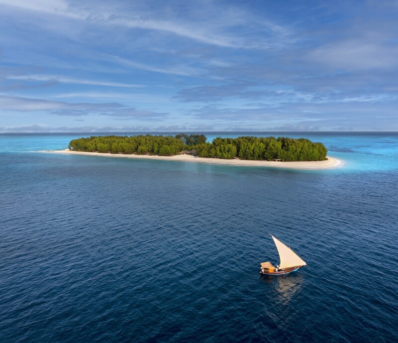 A small private island with white sand beaches and deep blue water surrounding it. A small sailboat is approaching it.
