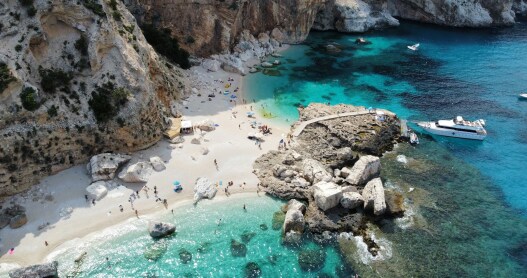 A white-sand beach alcove surrounded by large rocks, boulders and cliffs with people sunbathing on the beach and a boat out on the turquoise water in Sardinia, Italy