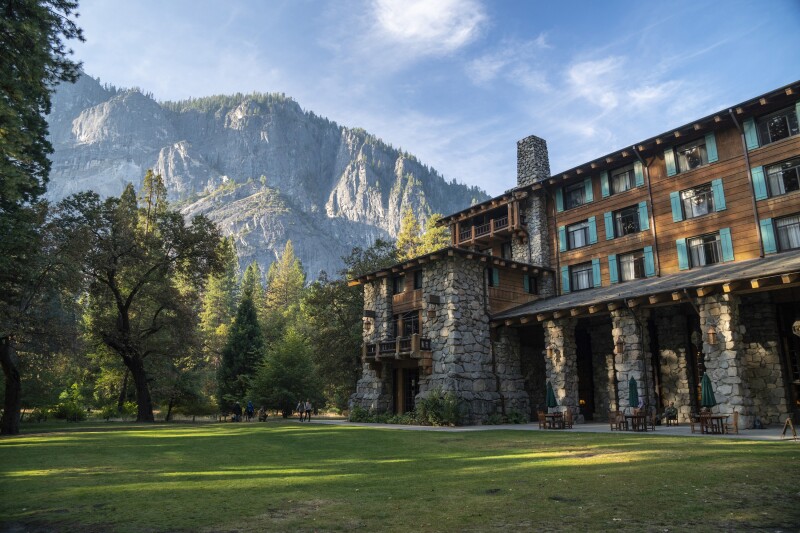 The Ahwahnee Hotel sits in the green grass valley of Yosemite, with a large mountain looming in the background. The hotel has a stone and wood exterior, with teal window shutters.