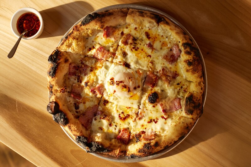 A white pizza on a wooden table, with a small container of chili oil next to it