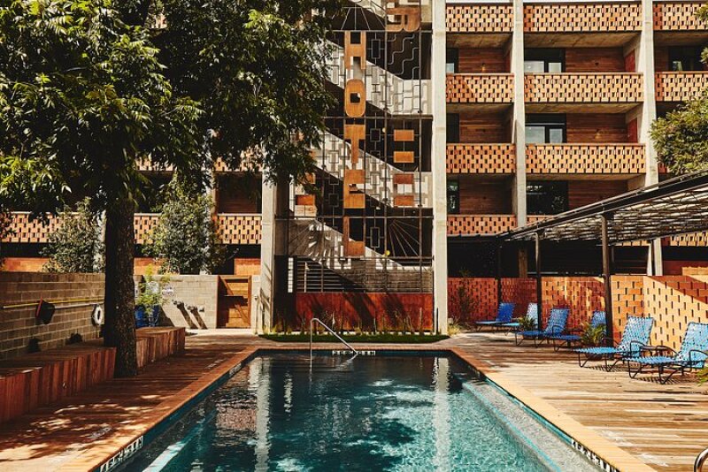 A long swimming pool outside the hotel, with lounge chairs and trees for shade. The "Hotel" sign is vertically formatted in brutalist font. The staircase can be seen from the outside and every room has a blalcony.