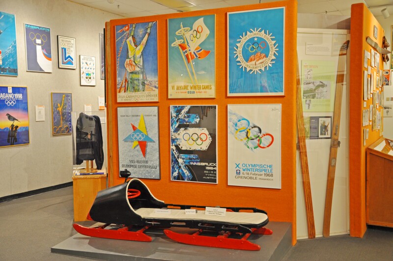Display of six Winter Olympics posters above a sled at Lake Placid Olympic Museum