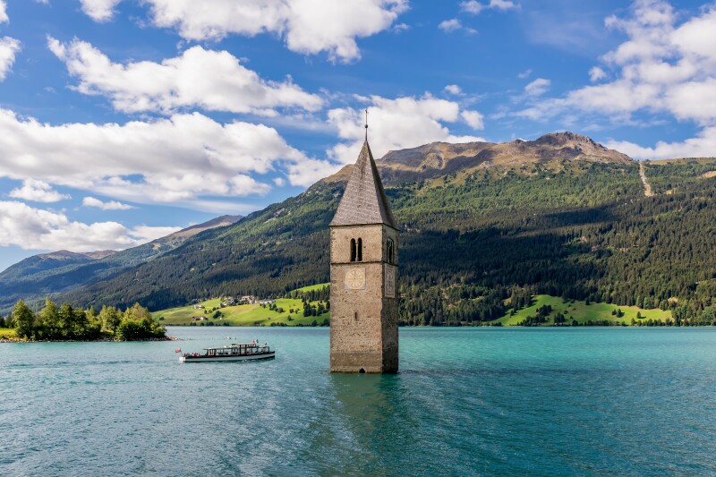 A bell tower surrounded by water with a boat floating past in a lake, with mountains in background