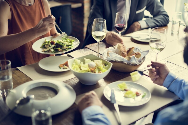One woman in dress and two men in suits at dining table with salads, bread, and wine