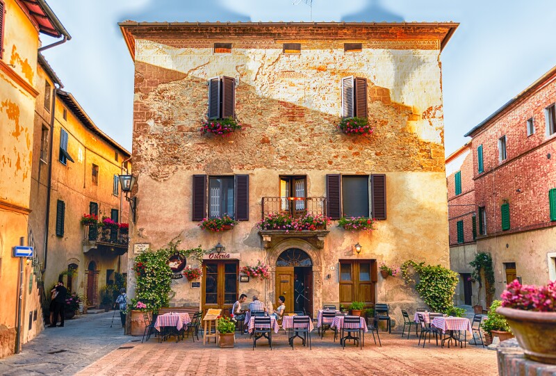 An old building in the medieval Tuscan village of Pienza with a restaurant and outdoor terrace with tables, chairs, and some patrons sitting outside on the ground floor
