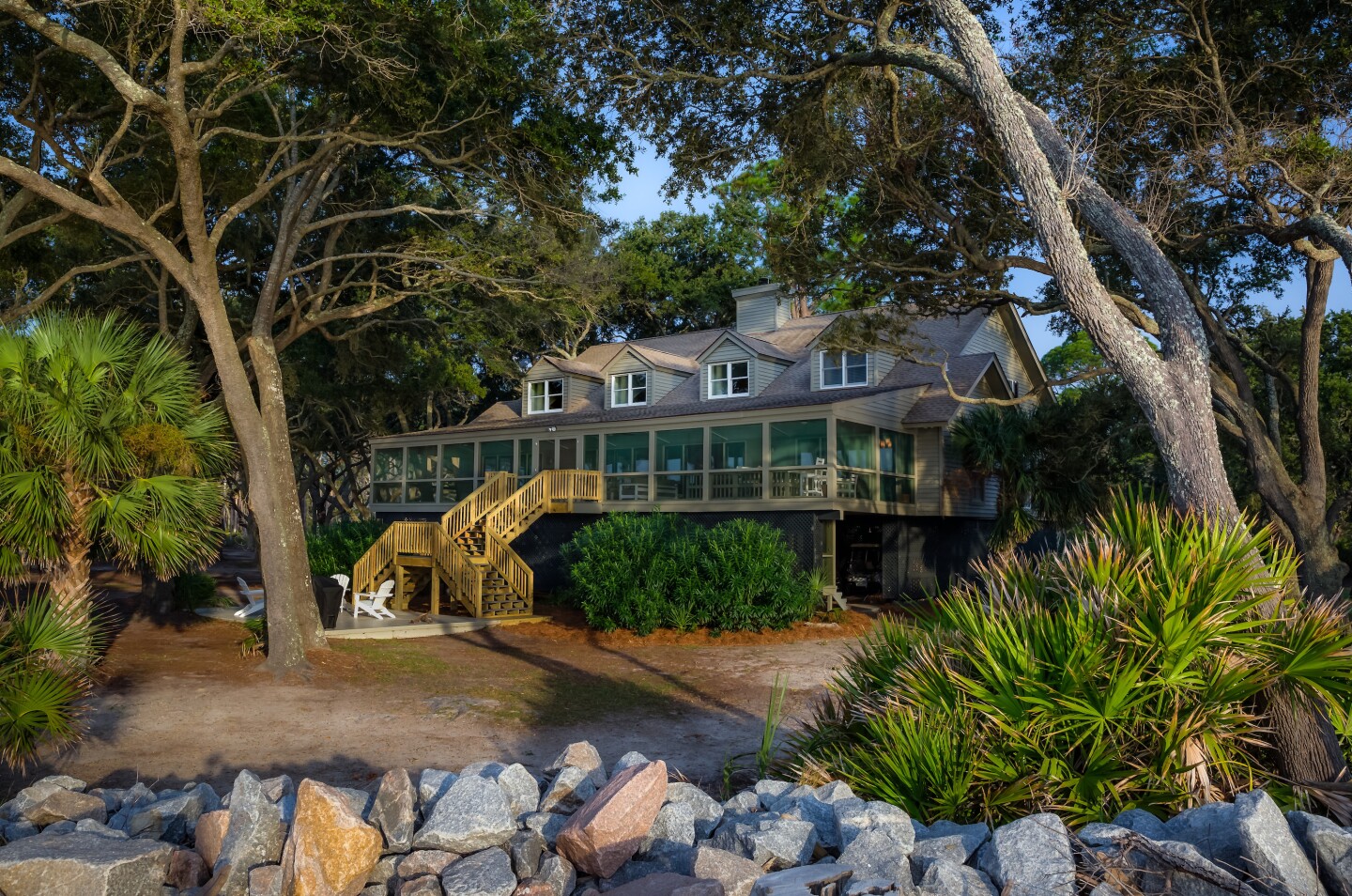 A large house with a wooden staircase and a screened porch, surrounded by trees and palmettos on a rocky shoreline