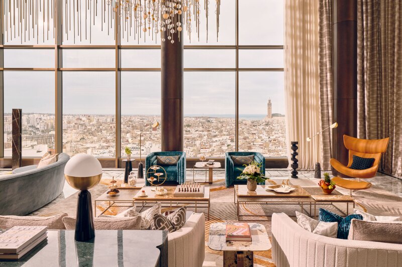 A room with pale pink furnishings, modernist chairs, globe lights, and a statement hanging light fixture; floor-to-ceiling windows overlook Moroccan cityscape